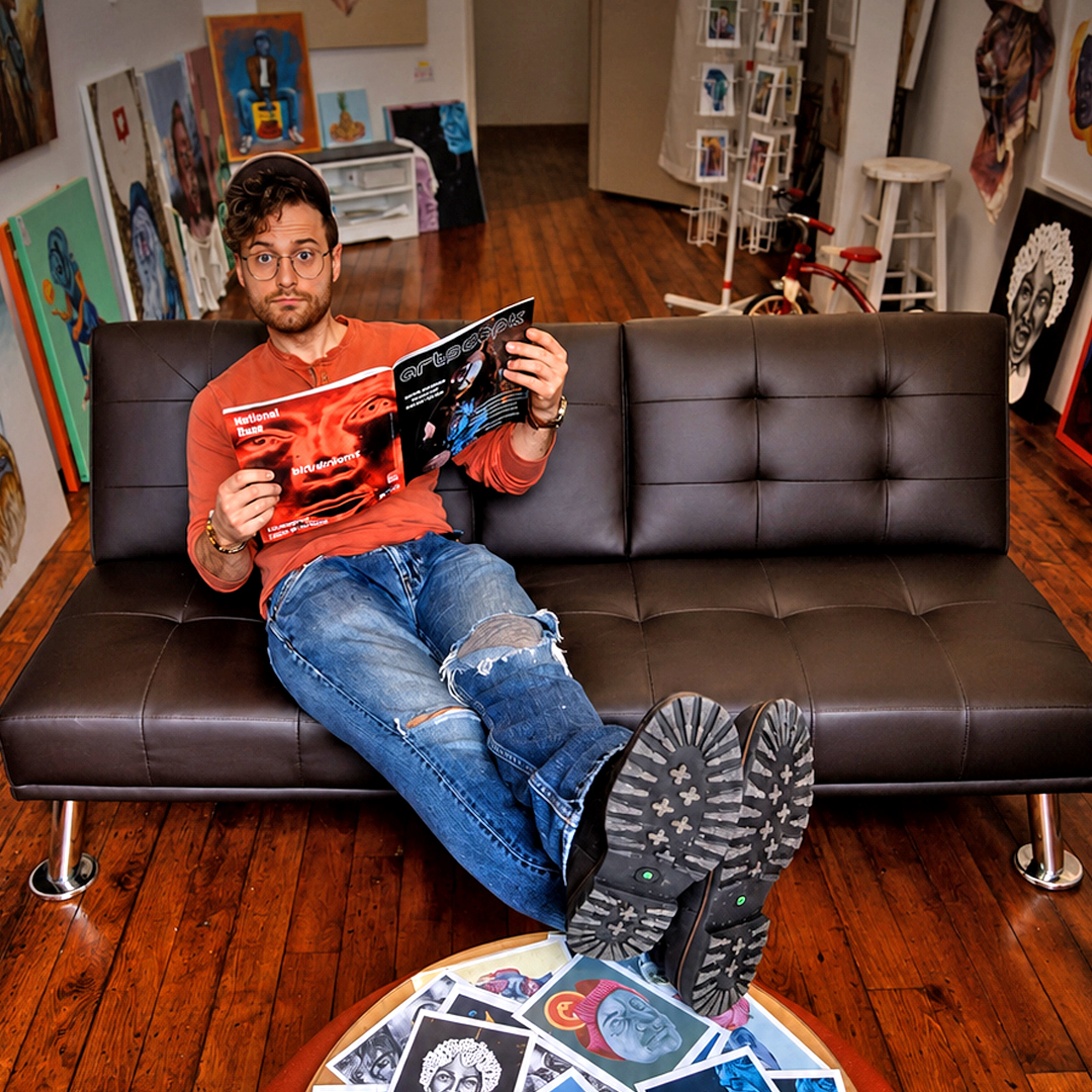 A man seated on a black sofa holding an art magazine, surrounded by paintings and prints.